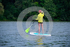 Athletic young girl-surfer riding on the stand-up paddle board in the clear waters of the on the background of green trees