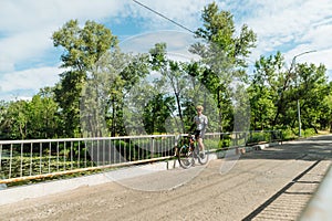Athletic young male cyclist stands in the park on the bridge and rests