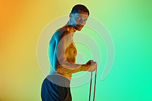 Athletic young man, with muscular, relief shirtless body training with resistance band against gradient blue yellow