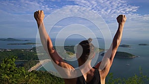 Athletic young man stands with his hands up on the edge of the mountain.