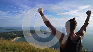 Athletic young man stands with his hands up on the edge of the mountain.