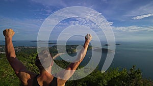 Athletic young man stands with his hands up on the edge of the mountain.