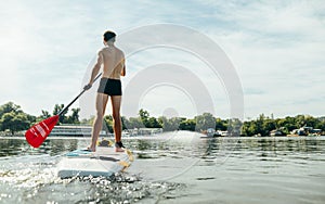 Athletic young man swims along the river on a paddle board, view from the back. Athlete trains on a sup board, actively paddles