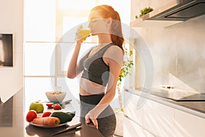 Athletic young red haired woman in the kitchen drinks a glass of fruit centrifuged juice