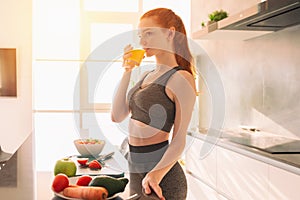 Athletic young red haired woman in the kitchen drinks a glass of fruit centrifuged juice