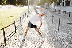 Athletic young tattooed man warming up by stretching before exercises outdoors, bearded fit guy in sportswear