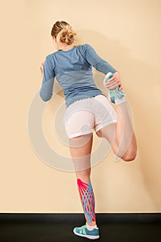 Athletic young woman with kinesio tape on her calf stretching her leg. Rear view. Kinesiology, physical therapy, rehabilitation.