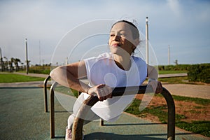 Athletic young woman in sportswear doing bar push-ups on cross bar, working out outdoors in the urban sportsground