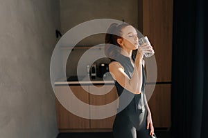 Athletic young woman wearing sportswear standing in home kitchen drinking fresh water after doing workout. Thirsty