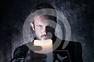 Atmospheric portrait of handsome knight with beard, looking at and holding candle up to camera while casting shadows