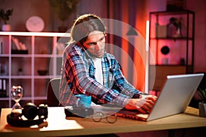 Attactive satisfied smiling young caucasian man in checkered shirt talking on phone and typing on laptop,