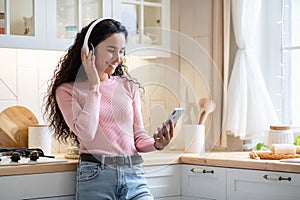 Attactive Smiling Lady With Smartphone And Wireless Headphones Listening Music In Kitchen