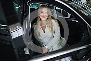 Attactive young bussines woman sitting in car in car showroom. Woman choosing new car. Beautiful blond hair female in