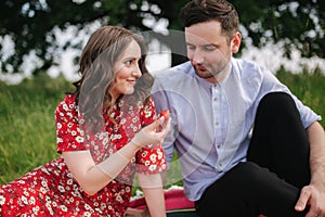 Attactive young woman with handsome man sits outdoors on red blanket. Woman give to man strawberry. Man taste berry