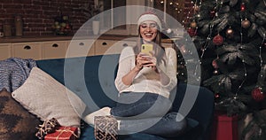 Attarctive Caucasian Blond Girl Wearing Santas Hat Looking Happy Sitting on a Sofa at Christmas Home Background Holding