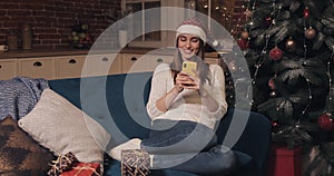 Attarctive Smiling Caucasian Blond Girl Wearing Santas Hat Looking Happy Sitting on a Sofa at Christmas Home Background