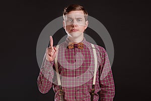 Attention! Strict Handsome young man in vintage shirt bow tie keeping finger raised and looking at camera while standing against