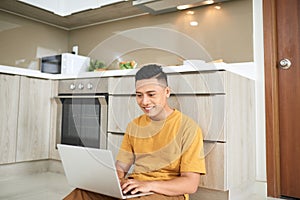 Attentive asian man using laptop while sitting on floor in kitchen