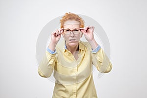 Attentive caucasian woman with short red hair looks scrupulously into distance, keeps hand on rim of spectacles.