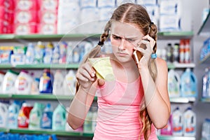 attentive girl standing in store choosing products while talking by phone and looking at shopping list