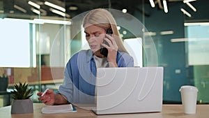 Attentive lady talking by mobile phone and making remarks in notebook by table