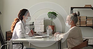Attentive middle aged older woman and young doctor in facial masks discussing treatment.