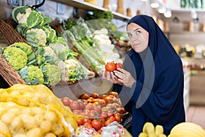 Attentive young Muslim woman purchaser choosing tomatoes in grocery store