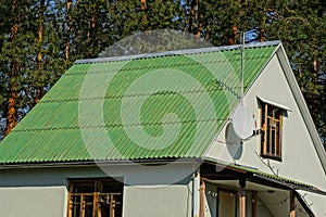 Attic of the house with windows and a roof under the green slate on the background of pines and sky