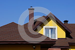 Attic of a yellow house with a window and a brown tiled roof against the sky
