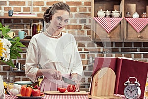 attractive adult housewife in vintage clothes cutting tomato and looking at camera