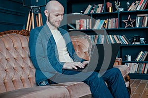 Attractive adult successful smiling bald man with beard in suit working at laptop on his rich cabinet