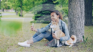 Attractive African American girl is taking selfie with cute dog resting in city park cuddling and caressing beautiful