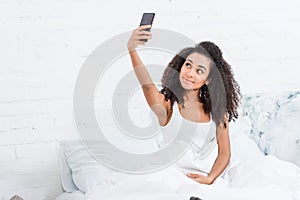 Attractive african american girl taking selfie on smartphone in bed during morning time