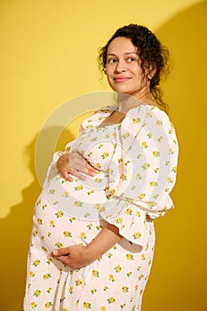 Attractive African American gravid woman, gently holding her belly, smiling looking aside, isolated yellow background