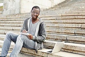 Attractive african-american student making notes sitting on stairs outdoors