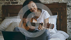 Attractive african american teenager girl concentraing learning to play guitar using laptop computer sitting on bed at