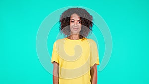 Attractive african american woman with afro hair in yellow basic clothing smiling to camera on teal blue wall studio