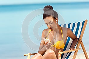 attractive african american woman laying on deck chair and listening music in earphones with smartphone in front of