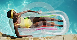 Attractive african american woman lying on inflatable holding drink and sunbathing in swimming pool