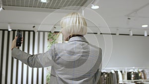 Attractive aged woman is taking a photo of herself at clothing store dressed in a grey suit, tracking shot, 360 degree