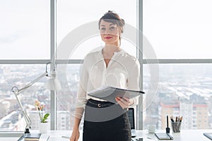 Attractive ambitious businesswoman standing in modern office, holding paper folder, looking at camera, smiling.