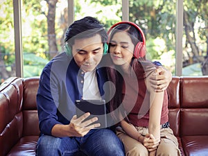 Attractive Asian couple wearing headphones sitting close together on couch in living room at home , man hugging woman , listening