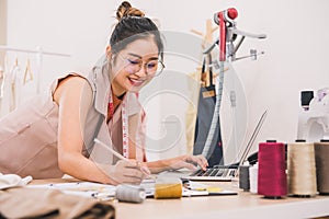 Attractive Asian female fashion designer working in home office workshop. Stylish fashionista woman creating new cloth design