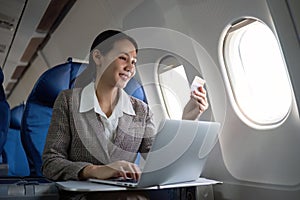 Attractive Asian female passenger on airplane sitting in comfortable seat using laptop and credit card, shopping using