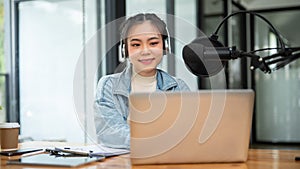 An attractive Asian female radio host is looking at her laptop screen, speaking into a microphone