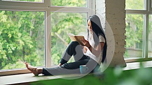 Attractive Asian girl student is reading book and smiling sitting on window ledge in modern apartment. Hobby, youth