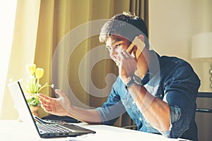 Attractive asian guy talking on phone while using laptop at workplace