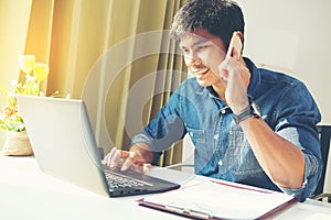 Attractive asian guy talking on phone while using laptop at workplace