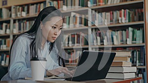 Attractive asian student girl sitting at table with pile of books in university library working on laptop computer