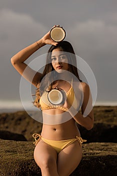 Attractive Asian woman holding coconut and wearing yellow swimsuit. Sexy woman sitting on the rock. Tanned skin. Summer concept.